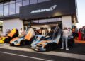 Three McLaren sports cars with butterfly doors open are displayed outside a McLaren dealership, drawing a crowd eager to witness this unique States of Endurance road trip event.