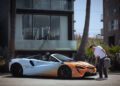 A man stands next to an orange and blue McLaren convertible sports car, holding a portable charging unit on a city street in front of modern buildings and palm trees—ready for his next States of Endurance road trip.