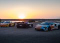 Three sports cars, including a McLaren, are parked on a dirt surface near water with a rock barrier and mountains in the background at sunset—capturing the spirit of an unforgettable States of Endurance road trip.