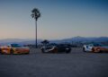 Three sports cars, including a sleek McLaren, are parked on a dirt lot at sunset, with a single palm tree and distant mountains setting the scene for an unforgettable road trip and ultimate States of Endurance.