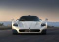 A white Maserati sports car, reminiscent of models seen at an MC12 auction, is parked on an open road at sunset, viewed from the front with mountains and buildings in the background.