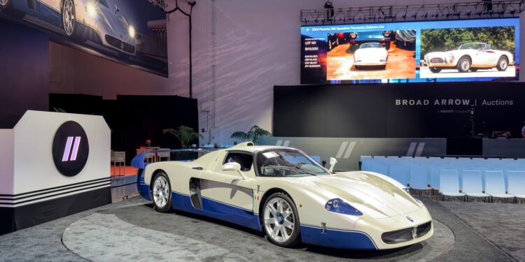 A record-breaking Maserati MC12 auction features a striking white and blue sports car showcased on a round platform, with empty seats and a large screen illuminating the background.
