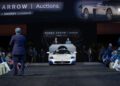 A record-breaking Maserati MC12 auction takes place as a white sports car is displayed on stage at a Broad Arrow event, with the auctioneer addressing attendees and two staff members standing behind the podium.