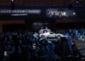 A stunning white and blue Maserati MC12 sports car is presented on stage at a crowded Broad Arrow auction, with a large banner and eager audience visible.