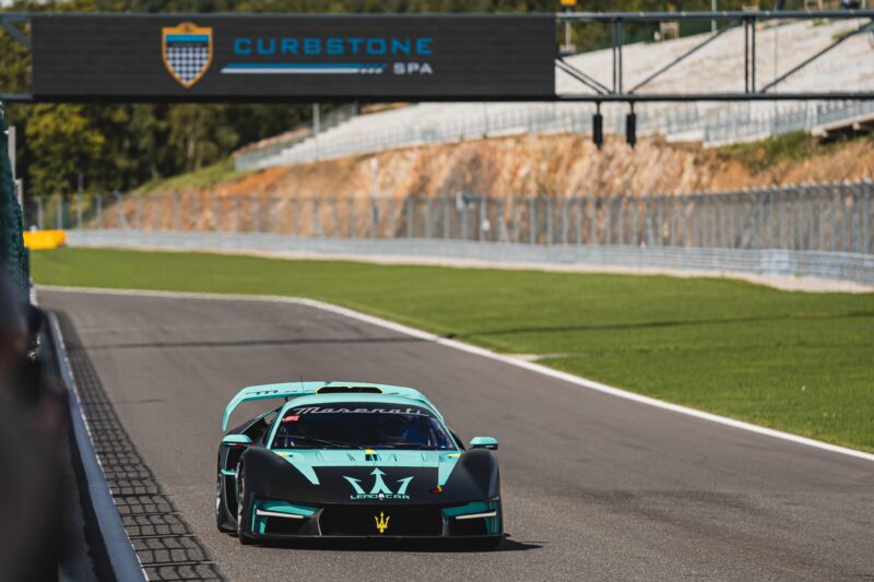 A turquoise Maserati MCXtrema race car speeds on a racetrack under a sign that reads "Curbstone at Spa," with barriers and green grass lining either side.
