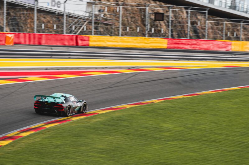A Maserati MCXtrema with a rear wing takes a corner on the Spa race track bordered by red and yellow curbs, with a fence and grandstands in the background.