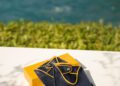A pair of navy blue leather driving gloves with yellow trim from the new collection rest on a yellow box, set on a white surface with the ocean and greenery in the background.