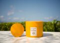A round yellow container and matching lid of Acqua di Parma’s new collection, placed on a white surface outdoors with greenery and blue sky in the background.