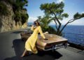 A woman in a yellow dress leans against a vintage Maserati convertible parked on a coastal road, with cliffs, trees, and the shimmering sea forming the perfect backdrop for the new collection.