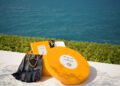 A round yellow Acqua di Parma box, black gloves, and a yellow booklet from the new collection are arranged on a white surface with greenery and the sea in the background.