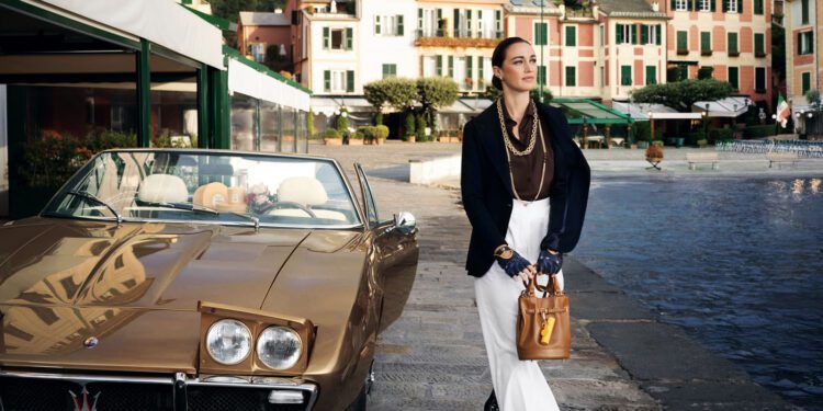 A woman walks beside a vintage brown Maserati parked by a waterfront, with colorful buildings and hills in the background, embodying the elegance of Acqua di Parma’s new collection.