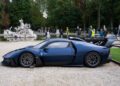 A blue Maserati MCXtrema sports car is parked on a gravel path near a fountain at the Turin Auto Show, with people and trees in the background.