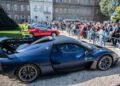 A matte blue Maserati MCXtrema is displayed outdoors at the Turin Auto Show, with a crowd of people and classic cars in the background.