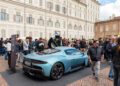 A crowd gathers around a blue Maserati sports car on display in a cobblestone plaza, surrounded by historic buildings at the Turin Auto Show.