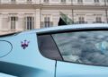 Close-up of the rear side of a light blue Maserati car, featuring the red Maserati trident logo and MCPURA badge, with a building reflected in the window—capturing the elegance often seen at the Turin Auto Show.