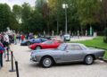 A classic silver coupe and a red Maserati sports car are displayed outdoors at the Turin Auto Show as people observe, with trees and statues in the background.