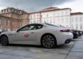 A silver Maserati MCPURA is parked on a stone courtyard in front of historic European buildings, reminiscent of scenes from the Turin Auto Show, with other cars lined up behind it.