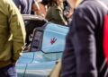 A close-up of a blue Maserati sports car at the Turin Auto Show, surrounded by people, with the iconic Maserati trident logo visible on the car's side pillar.