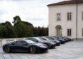 A row of Maserati sports cars is parked on a stone driveway beside a large, light-colored building, reminiscent of an exclusive Turin Auto Show, with trees in the background.