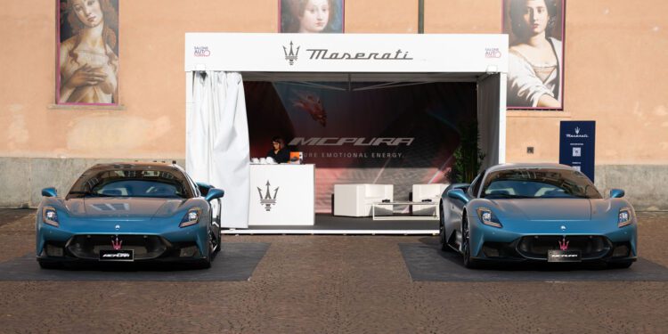 Two blue Maserati MC20 cars are parked in front of a Maserati display booth at the Turin Auto Show, with a person standing inside; two classical paintings adorn the peach-colored wall behind.