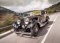 A classic black Rolls-Royce Phantom glides along a winding road, with rolling hills and charming houses in the background—capturing the spirit of Goodwood Revival.