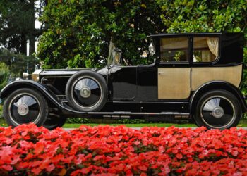 A vintage black and beige Rolls-Royce is parked on a road, with bright red flowers in the foreground and green trees in the background – a scene reminiscent of the elegance found at Goodwood Revival.