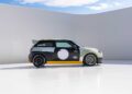 A modified Mini Cooper, inspired by MINI’s Rally DNA Gets a Deus Makeover in Two Radical JCW Builds, sports racing decals and a large rear spoiler as it sits parked on a smooth white surface against a curved white backdrop and partly cloudy sky.