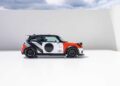 A compact car with a bold black, white, and orange geometric design is parked on a flat white surface against a curved white wall and cloudy sky, echoing MINI’s Rally DNA Gets a Deus Makeover in Two Radical JCW Builds.