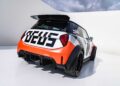 A compact race car with wide tires, large rear spoiler, and bold "DEUS" lettering on the back showcases MINI’s Rally DNA Gets a Deus Makeover in Two Radical JCW Builds, parked on a white surface against a cloudy sky.