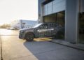 A camouflaged BMW iX5 Hydrogen prototype car is partially exiting a garage in an industrial area during daylight, hinting at the future of hydrogen and series production vehicles.