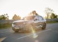 A camouflaged BMW iX5 Hydrogen prototype SUV drives on a road with trees and a metal barrier in the background on a sunny day.
