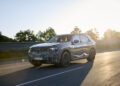 A camouflaged SUV-style test vehicle, possibly the new BMW iX5 Hydrogen, drives on a road at sunset, with trees and a metal barrier visible in the background.