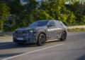 A camouflaged BMW iX5 Hydrogen test vehicle drives on a road, surrounded by greenery and trees in the background.