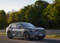 A camouflaged BMW iX5 Hydrogen SUV drives on a road, with trees and a guardrail visible in the background, showcasing the future of hydrogen mobility ahead of potential series production.