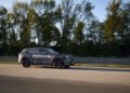 A camouflaged BMW iX5 Hydrogen vehicle drives on a road lined with trees and a metal barrier, showcasing the future of hydrogen mobility ahead of series production.
