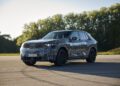 A BMW iX5 Hydrogen SUV test vehicle with camouflage wrap and “BMW Hydrogen” written on the side is parked on a road, hinting at future series production, with trees and blue sky in the background.