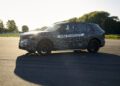 A camouflaged BMW iX5 Hydrogen test vehicle labeled "BMW HYDROGEN" is parked on an open paved area with trees in the background, showcasing the brand’s commitment to hydrogen car innovation ahead of potential series production.