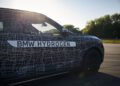 A camouflaged BMW iX5 Hydrogen, labeled "BMW HYDROGEN," is parked on a road with trees and clear sky in the background, highlighting the brand’s step toward series production of innovative hydrogen vehicles.