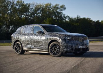 A BMW iX5 SUV test vehicle with a camouflage wrap and “BMW Hydrogen” label is parked on a paved surface in daylight, hinting at hydrogen Series Production, with trees in the background.