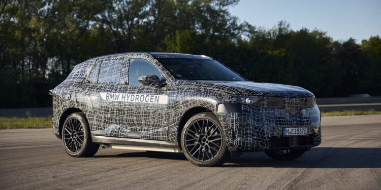 A BMW iX5 SUV test vehicle with a camouflage wrap and “BMW Hydrogen” label is parked on a paved surface in daylight, hinting at hydrogen Series Production, with trees in the background.