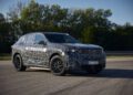 A camouflaged BMW iX5 SUV test vehicle labeled "BMW HYDROGEN" is parked on a paved surface with trees in the background, hinting at future Series Production of hydrogen-powered models.