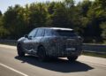 A BMW iX5 Hydrogen prototype SUV with "BMW Hydrogen" branding drives on a road, featuring camouflage wrap and a partially visible license plate. This hydrogen vehicle is captured with trees in the background.