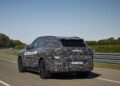A camouflaged BMW iX5 Hydrogen vehicle drives on a paved road with trees and clear sky in the background, showcasing the future of hydrogen vehicle technology ahead of potential series production.