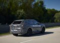 A camouflaged BMW iX5 Hydrogen SUV prototype drives on a road, surrounded by greenery under a clear blue sky, hinting at the brand's potential steps toward series production.