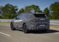 A camouflaged BMW iX5 Hydrogen test vehicle drives on a road with trees and a metal barrier in the background, highlighting BMW’s commitment to hydrogen technology ahead of potential series production.