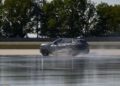 A BMW iX5 Hydrogen SUV drives through water on a test track, creating a dramatic spray with trees in the background, highlighting the innovation approaching series production.