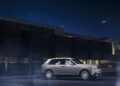 A silver Rolls-Royce Cullinan SUV is parked at night in front of a modern building, its Starlight Headliner mirroring the starry sky above.