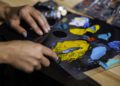 A person mixes yellow and blue oil paint on a palette with a palette knife, surrounded by various paint colors and tubes on a wooden table, inspired by the cosmic elegance of the Starlight Headliner found in the Rolls-Royce Cullinan.