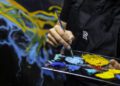 A person in a black apron holds a brush and palette with blue, yellow, and turquoise paint, creating a colorful abstract painting inspired by the Cosmos in the background.