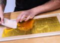 A person applies gold leaf sheets to a rectangular surface using a thin paper sheet, on a wooden table.